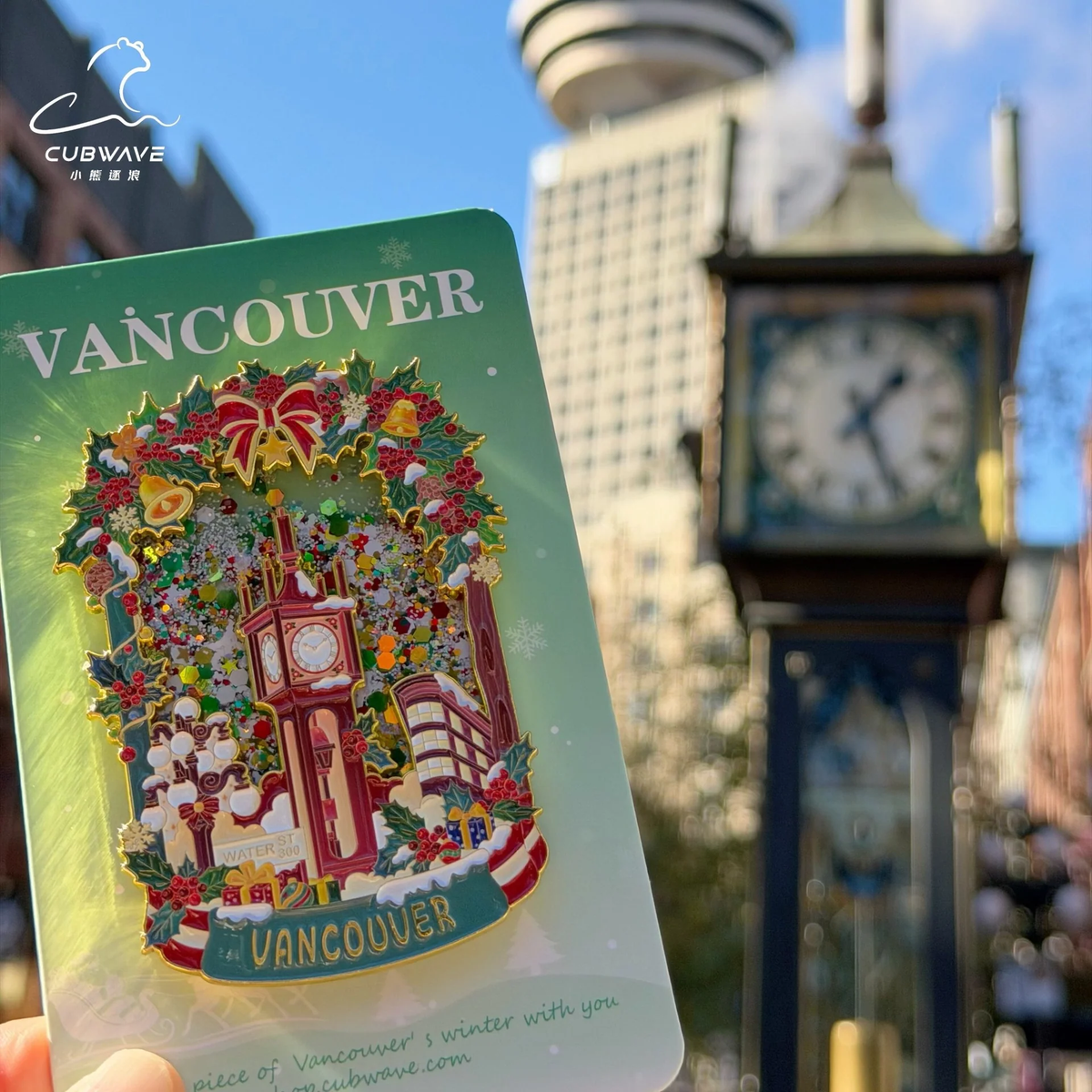 Cubwave Vancouver Christmas Steam Clock snow-flow fridge magnet on green packaging, with holly, bells and a red bow, photographed in front of Gastown’s Steam Clock.