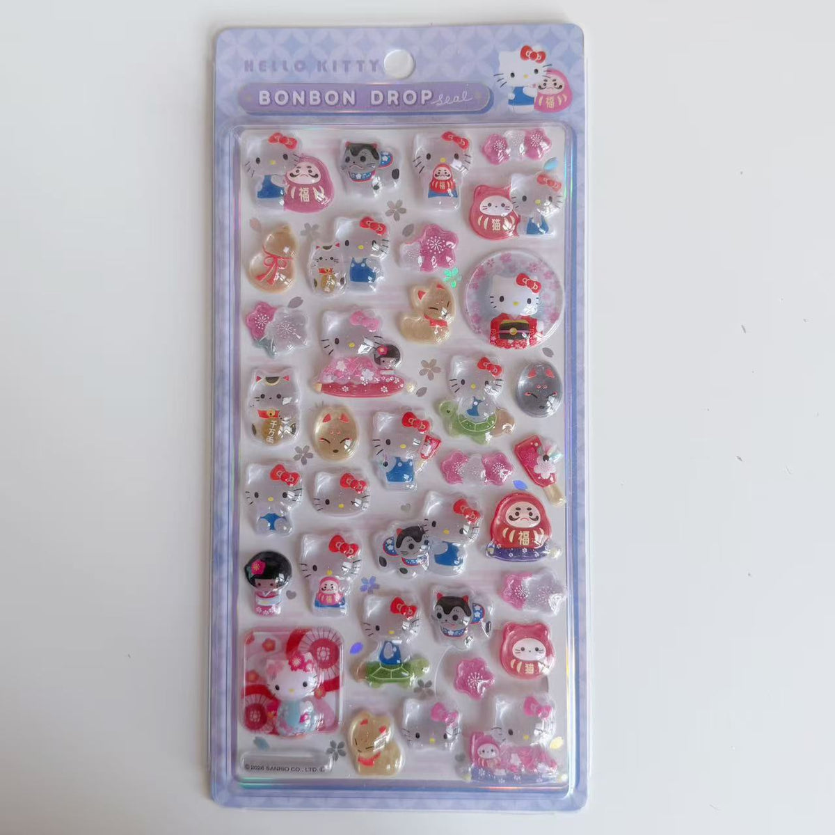 Hello Kitty BONBON DROP Lucky Charms 3D sticker sheet in purple packaging with daruma dolls, beckoning cats and cherry blossoms.